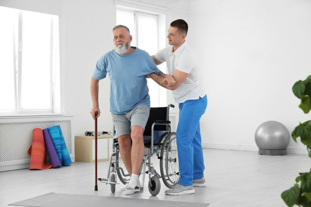 Ergotherapie Vaihingen: Intensivtraining Physiotherapeut hilft älterem Mann aus dem Rollstuhl für Gehtraining